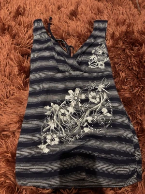 Randig blommig topp med knytdetalj  - Supersöt kortklänning i marinblått och silver med smala ränder och blommigt tryck framtill. Klänningen har v-ringning, knytband i ryggen och är ärmlös. Materialet känns mjukt och stretchigt, perfekt för sommarens alla dagar❤️❤️