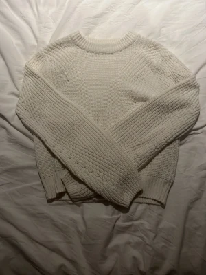 Vit stickad tröja med strukturmönster - Mysig vit stickad tröja med grov struktur och rund hals. Tröjan har långa ärmar och ribbade muddar vid ärmslut och nederkant. Perfekt för kyliga dagar och enkel att matcha med jeans eller kjol.