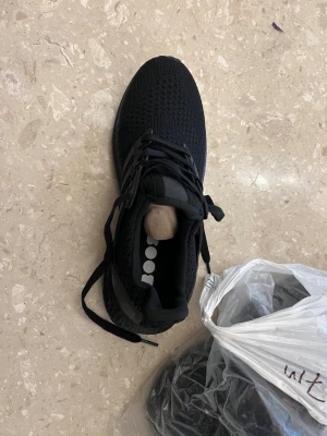 Svarta Adidas Ultraboost sneakers - Säljer ett par helsvarta Adidas Ultraboost sneakers med stickad ovandel och klassiska tre ränder på sidan. Skorna har snörning, platt sula och en bekväm Boost-mellansula. Perfekta för dig som gillar stilrena och sportiga skor.