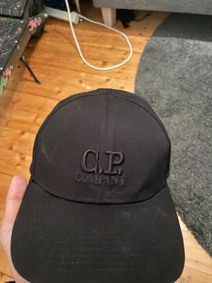 Svart keps från C.P. Company - Säljer en stilren svart keps från C.P. Company med broderad logga framtill och justerbart spänne bak. Kepsen är tillverkad i 100% bomull och har en klassisk form som passar till det mesta.