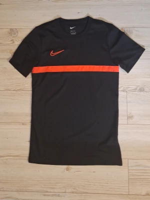 Svart Nike Dri-Fit t-shirt med röd detalj - Snygg svart Nike Dri-Fit t-shirt med en röd rand över bröstet och röd Nike-logga. Tillverkad i ett lätt och ventilerande funktionsmaterial som passar perfekt till träning eller sport. Klassisk rund hals och korta ärmar för en sportig look.