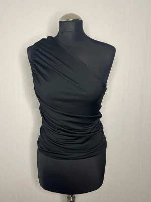 Svart oneshoulder topp från H&M - Snygg svart oneshoulder topp från H&M i storlek S. Toppen har draperad effekt och sitter tight mot kroppen. Tillverkad i mjukt och stretchigt material som ger en smickrande siluett. Perfekt för dig som vill ha en trendig och stilren look.