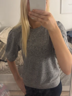 Grå stickad topp från 157 - Mysig grå stickad topp från 157 med rund halsringning och puffiga korta ärmar. Enkel och stilren design som passar perfekt till lågmodjade jeans.