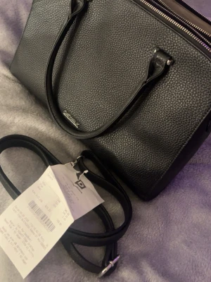 Svart handväska från Catwalk - Säljer en stilren svart handväska från Catwalk präglat skinn. Väskan har två handtag, avtagbar axelrem och flera fack med dragkedja för smart organisering. Perfekt storlek för allt du behöver ha med dig och snygg metallogo framtill. Mindre dator och skrivblock får plats. Perfekt till skola/jobb. Liten vit fläck.