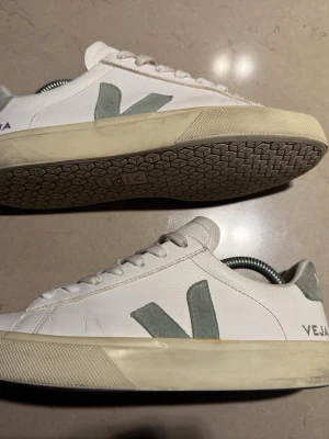 Veja Skor - Snygga vita sneakers från Veja med grönt V på sidan och beige sula. Skorna har snörning och är tillverkade i skinn med detaljer i mocka. Perfekta för dig som gillar stilrena och klassiska sneakers med en twist.