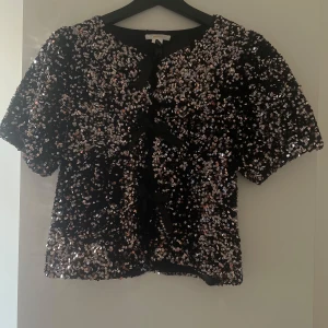 Svart glittrig blus med paljetter - Glittrig svart blus från Gina tricot endast använd en gång! Den är täckt med silvriga paljetter som ger maxad partykänsla. Kortärmad modell med tre svarta rosettknutar framtill. Perfekt för dig som vill sticka ut och glänsa på festen!🩶