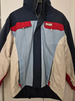 Vinterjacka/Skidjacka - Säljer en retro vinterjacka från Gianiferoti Outdoor Wear i färgerna marinblå, ljusblå, röd och beige. Jackan har huva, dragkedja, snörning i nederkant och coola färgblock. Perfekt för dig som gillar vintage och streetwear.