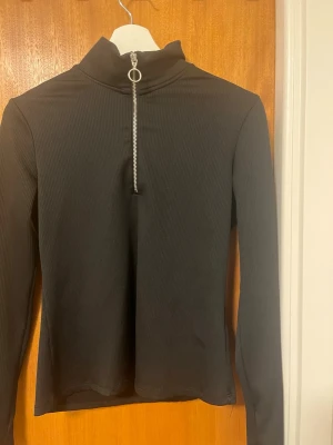 Svart ribbad polotopp med dragkedja - Säljer en svart ribbad polotopp med lång ärm och en snygg silverfärgad dragkedja framtill med ringdetalj. Toppen är i stretchigt material och har en tight passform som sitter snyggt på kroppen. Perfekt att styla med jeans eller kjol. Materialett är jätte könt och mjukt.