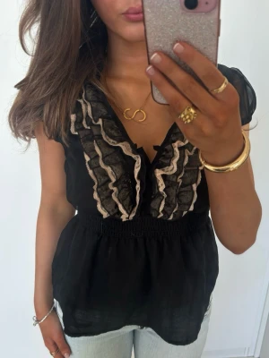 Svart blus med volang och spets - Säljer en svart blus med transparenta korta ärmar och v-ringning. Framtill har den dekorativa volanger och spetsdetaljer i beige längs urringningen. Jag är 168 och det är mina bilder, köp via köp nu direkt eller Swish, kvar tills markerad som såld 💖 