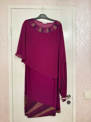 Lila långklänning med glittriga detaljer  - Köpt utomlands för ett par år sedan. Endast använd en gång. Långklänning i flowy material med glittriga pärldetaljer runt halsringningen och längst ner. Asymmetrisk transparent överdel. Två delar. Snyggt mönster i guld och lila nedtill. Perfekt för festliga tillfällen. Då den är köpt utomlands är storleken på lappen inte i EU, men den passar perfekt för min mamma som har storlek L och bröstmåttet är 96cm (48cm x 2).