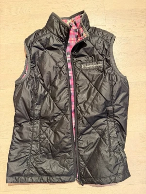 Svart och rutig dunväst Peak Performance (vändbar) - Säljer en vändbar dunväst från Peak Performance i storlek L. Ena sidan är svart och quiltad med dragkedja, andra sidan har ett rosa, vitt och svart rutigt mönster. Västen har hög krage och fickor på svarta sidan. Perfekt för dig som vill ha två stilar i en.
