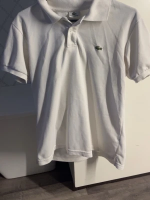 Vit pikétröja från Lacoste - Klassisk vit pikétröja från Lacoste med korta ärmar och krage. Tröjan har knappar framtill och den ikoniska gröna krokodilloggan broderad på bröstet. Tillverkad i bomull för en skön och luftig känsla.