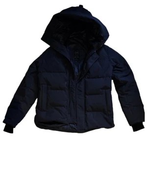 Canada Goose Macmillan - Säljer en svart pufferjacka från Canada Goose med huva och diskret logga på ärmen. Jackan har dragkedja framtill, två stora fickor och ribbade muddar vid ärmsluten. Perfekt för kalla dagar och riktigt snygg passform. Canada goose macmillan parka black label 