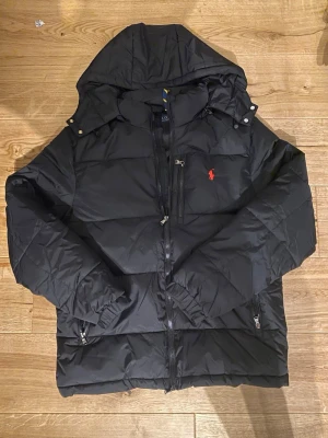 Svart pufferjacka från Polo Ralph Lauren - Säljer en svart pufferjacka från Polo Ralph Lauren med huva och dragkedjor. Jackan har det klassiska röda Polo-logot på bröstet och flera praktiska fickor med dragkedja. Perfekt för kalla dagar och riktigt snygg till streetwear-stilen.
