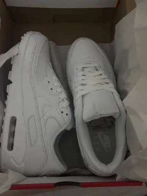 Vita Nike Air Max sneakers - Säljer ett par helvita Nike Air Max sneakers med klassisk chunky sula och synlig luftbubbla. Skorna har snörning och är tillverkade i skinn och syntetmaterial. Perfekta för dig som vill ha en clean och stilren look.