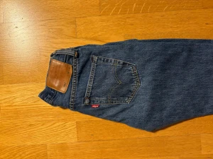Levi's 501 blå jeans straight fit - Klassiska Levi's 501 jeans i blå tvätt med raka ben och normal passform. Jeansen har fem fickor, knappgylf och den ikoniska bruna läderpatchen bak i midjan. Tillverkade i slitstarkt bomullsjeans med kontrastsömmar och röd Levi's-tag på bakfickan.