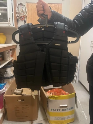 Svart skyddsväst från Jacson - Säljer en svart skyddsväst från Jacson med quiltad design och breda kardborreband i sidan för justerbar passform. Västen är vadderad och har en sportig look, perfekt för ridning. Passar dig som vill ha både säkerhet och stil. Passar ungefär för 10-11 år.