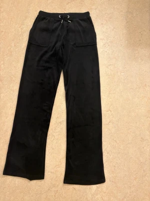 Svarta mjukisbyxor Juicy Couture - Svarta mjukisbyxor från Juicy Couture med bred resårmidja och snörning. Byxorna har raka ben, stora fickor fram och bak med lock och metallknappar med logga. Materialet på byxorna är velour.