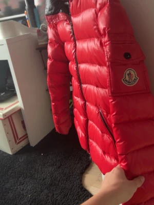 Röd Moncler pufferjacka - Säljer en riktigt snygg röd pufferjacka från Moncler. Jackan har svart innerfoder, två dragkedjeförsedda fickor och klassisk Moncler-logga på ärmen. Fylld med dun för maxad värme och har en hög krage. Perfekt för kalla dagar och riktigt stilren.