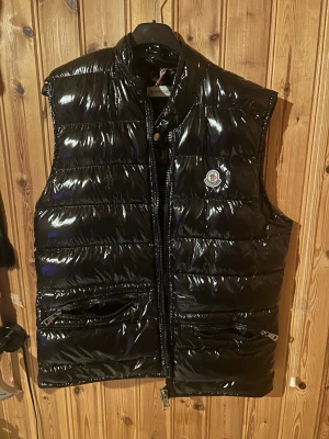 Svart dunväst från Moncler - Säljer en glansig svart dunväst från Moncler med hög krage och två dragkedjefickor framtill. Västen har quiltad design och klassisk Moncler-logga på bröstet. Perfekt att slänga över en hoodie för en cool look.