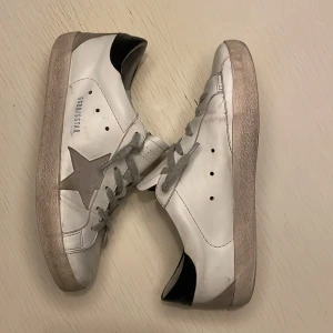 Golden Goose Superstar sneakers vit/svart - Golden Goose Superstar sneakers i vitt skinn med svart häl och grå stjärndetalj på sidan. Skorna har grå snörning, platt sula och klassisk rund tå. Snygg kontrast mellan det vita och svarta samt den ikoniska stjärnan på sidan. Säljer pågrund av att de har blivit försmå