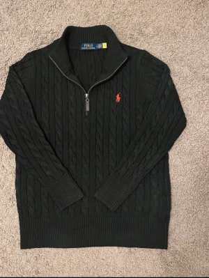 Svart kabelstickad tröja Polo Ralph Lauren - Kabelstickad svart tröja från Polo Ralph Lauren med röd broderad logga på bröstet. Tröjan har hög krage och half zip-dragkedja. Ribbstickade muddar vid ärmslut och nederkant. Perfekt för dig som gillar klassisk och stilren look.