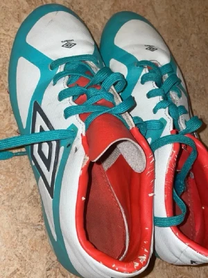Turkosa och vita Umbro fotbollsskor - Säljer ett par fotbollsskor från Umbro i turkos och vitt med röda detaljer. Skorna har snörning och en klassisk Umbro-logga på sidan. Insidan är röd och materialet är syntet, perfekt för dig som vill sticka ut på planen.