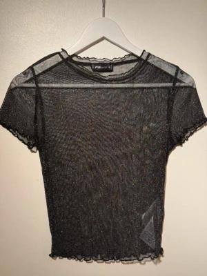 Glittrig mesh topp från F.A. Sister - Säljer en svart, glittrig mesh topp från F.A. Sister. Toppen är genomskinlig med korta ärmar och har vågiga kanter vid ärmslut och nederkant. Perfekt att styla lager på lager eller till festliga tillfällen.