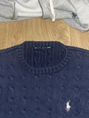 Mörkblå stickad tröja från Ralph Lauren - Säljer en mörkblå stickad tröja från Ralph Lauren med klassiskt kabelstickat mönster och rund hals. Tröjan har lång ärm och den ikoniska vita loggan broderad på bröstet. Perfekt för dig som vill ha en stilren och tidlös look.