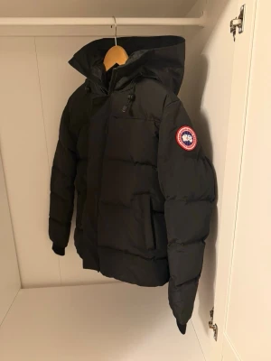 Svart dunjacka  - Säljer en svart dunjacka med huva och klassisk logga på ärmen. Jackan har dragkedja, knappar och justerbara snören vid huvan. Perfekt för kalla dagar och riktigt varm. Passar dig som vill ha en stilren och funktionell vinterjacka.