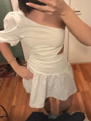 Vit one shoulder kortklänning - Säljer en vit kortklänning med one shoulder-design och puffärm. Klänningen har en snygg cut-out i midjan och volang nedtill. Tillverkad i ett lätt bomullsliknande material, perfekt för sommarens alla tillfällen. Perfekt inför studenten 
