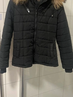 Svart pufferjacka med pälls från Belle - Säljer en svart pufferjacka från Belle i storlek XS. Jackan har quiltad design, dragkedja framtill och två fickor. Huva med fluffig fuskpäls i beige/brun ton. Perfekt för kalla dagar och riktigt snygg till alla outfits. Passar en S också. Köpt för 1200kr