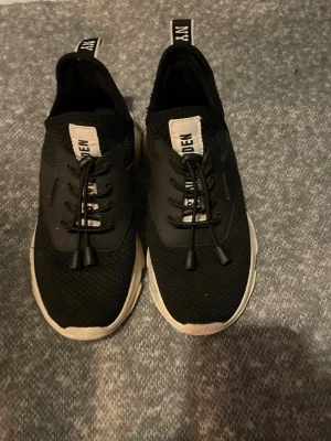 Svarta sneakers från Steve Madden - Säljer ett par svarta sneakers från Steve Madden med vit sula och elastisk snörning. Ovandelen är i mesh-liknande syntetmaterial och har en vit detalj med logga på plösen. Skorna har rund tå och platt sula för skön komfort.
