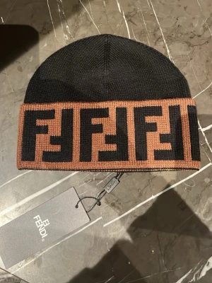 Fendi svart och brun mössa - Säljer en snygg svart mössa från Fendi med bred brun kant och ikoniskt FF-mönster i svart. Mössan är ribbstickad och har en klassisk passform. Perfekt accessoar för dig som vill ha något lyxigt och trendigt till vintern.