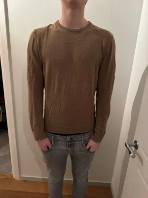 Beige stickad tröja med rund hals - En enkel och stilren beige stickad tröja med rund halsringning och långa ärmar. Tröjan har ribbade muddar vid ärmslut och nederkant, vilket ger en snygg passform. Perfekt att matcha med jeans för en clean look.