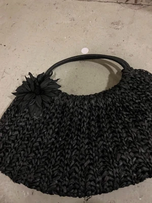 Svart handväska med blomdetalj - Säljer en svart handväska i flätad syntet med rundade handtag och en stor dekorativ blomma på sidan. Väskan har ett rymligt fack med tryckknapp och är perfekt för dig som vill ha en unik accessoar. Passar till många olika stilar och outfits.