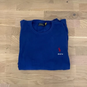 Blå stickad tröja från Polo Ralph Lauren - Säljer en klassisk blå stickad tröja från Polo Ralph Lauren med rund hals och den ikoniska röda loggan broderad på bröstet. Tröjan har långa ärmar och ribbade muddar vid ärmslut och nederkant. Perfekt för dig som gillar stilrena plagg. Strlk är L men är mer L/M