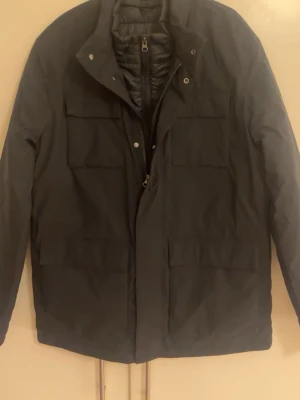 Svart jacka från John Henric - Säljer en svart jacka med hög krage och fyra stora fickor framtill. Jackan har dragkedja och tryckknappar, samt quiltad insida för extra värme. Perfekt för kyligare dagar och har en stilren, enkel look som passar till det mesta. Pris kan diskuteras. Ordinarie pris 2550kr