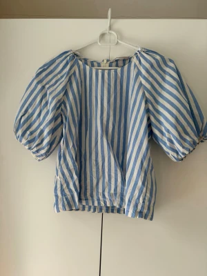 Blårandig puffärmad blus - Säljer en blus med blå och vita vertikala ränder och stora puffärmar. Toppen har en lös passform och är tillverkad i ett lätt bomullstyg, perfekt för varma dagar. Den har rund halsringning och avslappnad siluett.