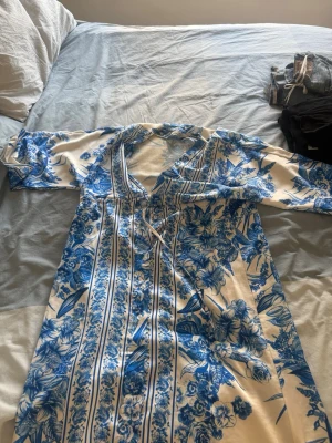 Blåvit blommig långklänning New Collection - Säljer en långklänning från New Collection i blått och vitt med blommigt och randigt mönster. Klänningen har v-ringning, knappar framtill och långa ärmar. Materialet känns mjukt och svalt, perfekt för varma dagar.