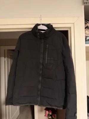 Svart pufferjacka från Porsche - Säljer en svart pufferjacka från Porsche med hög krage, diskret Porsche-logga på bröstet och patch på ärmen. Jackan har dragkedja framtill, bröstficka med dragkedja och två sidofickor. Perfekt för kalla dagar och riktigt snygg streetstil.