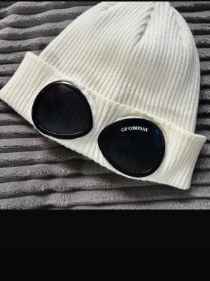 Vit ribbad mössa från C.P. Company - Säljer en vit ribbad mössa från C.P. Company med ikoniska svarta glasögonlinser framtill. Mössan är stickad och har en uppvikt kant för extra stilpoäng. Perfekt accessoar för dig som vill sticka ut och hålla dig varm.