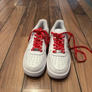 Nike Air Force 1 x Supreme vita sneakers - Vita Nike Air Force 1 sneakers i skinn med röda Supreme-skosnören knappt använda i princip helt nya.. Klassisk siluett med perforerad tå, platt sula och rund tå. Snygg kontrast mellan det vita och de röda detaljerna. Perfekta för dig som gillar streetwear och vill sticka ut. 