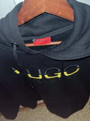 Hugo boss hoodie  - Snygg Hugo Boss hoodie i svart med en exklusiv guldig och svart design/logga på framsidan. Hoodien är i storlek S och i som nytt skick, perfekt för dig som vill ha en stilren och lyxig vardagslook.  ✨ Egenskaper: 	•	Märke: Hugo Boss 	•	Färg: Svart 	•	Storlek: S 	•	Skick: Som ny