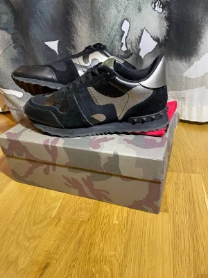 Valentino Garavani camo sneakers - Säljer ett par svarta och silverfärgade Valentino Garavani sneakers med camomönster och coola nitar på sulan. Skorna har snörning, rund tå och är tillverkade i en mix av mocka, skinn och textil. Perfekta för dig som vill sticka ut med exklusiva detaljer.