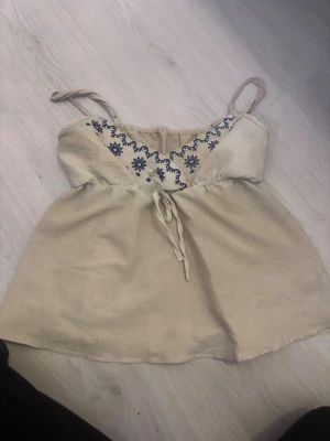 Beige broderad linnetopp från SHEIN - Säljer en beige linnetopp från SHEIN i storlek S. Toppen har tunna axelband och är dekorerad med blå broderade blommor och detaljer framtill. Materialet är lätt och luftigt, perfekt för varma dagar. Söt volangkant upptill ger en extra touch.