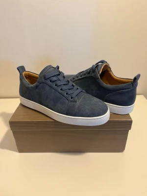 Blå mockasneakers från Louboutin - Snygga blå sneakers från Christian Louboutin i mjuk mocka med klassisk röd sula och vita kanter. Skorna har rund tå, snörning och beige innersula med guldfärgad logga. Perfekta för dig som vill ha en exklusiv touch på din outfit.