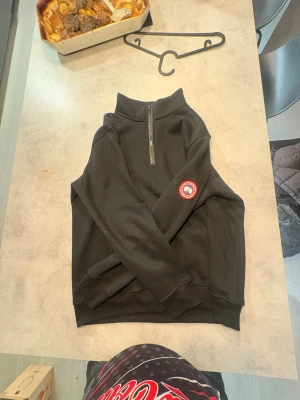 Canada Goose Quarter Zip - Svart långärmad tröja från Canada Goose med half zip och klassisk logga på ärmen. Tröjan har en hög krage och är tillverkad i mjukt material som passar perfekt till kyligare dagar. Enkel och stilren design som funkar till det mesta.