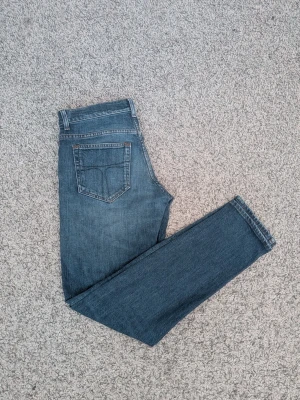 Tiger of Sweden jeans - Säljer ett par klassiska blå jeans från tiger of Sweden med raka ben och slim passform. Jeansen har fem fickor, snygga kontrastsömmar och ett brunt läderpatch bak i midjan. Tillverkade i slitstarkt denimtyg som ger en tidlös look. Storlek W29 L32. Hör av er vid frågor!