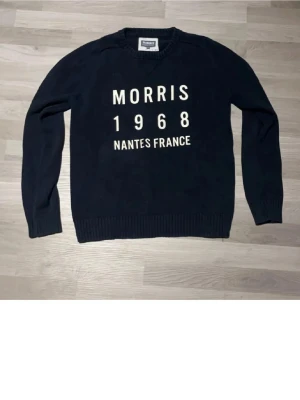 Mörkblå stickad tröja från Morris - Säljer en mörkblå stickad tröja från Morris med vit text framtill: 'Morris 1968 Nantes France'. Tröjan har rund halsringning, ribbade muddar och lång ärm. Perfekt för dig som gillar klassisk och stilren design.